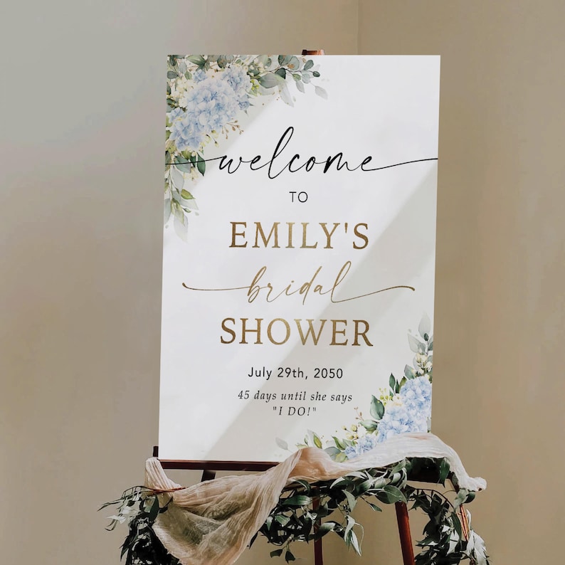 May include: A white sign with the words "welcome to Emily's bridal shower" in elegant script. The sign is decorated with blue floral accents and greenery. The date July 29th, 2050, is displayed, along with the text "45 days until she says 'I DO!'"