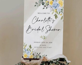 May include: A white sign with the text "WELCOME TO Charlotte's Bridal Shower" in elegant script. Floral accents in yellow and blue adorn the top corners. The sign also includes the date, "SUNDAY, JUNE 20, 2050," and countdown details.