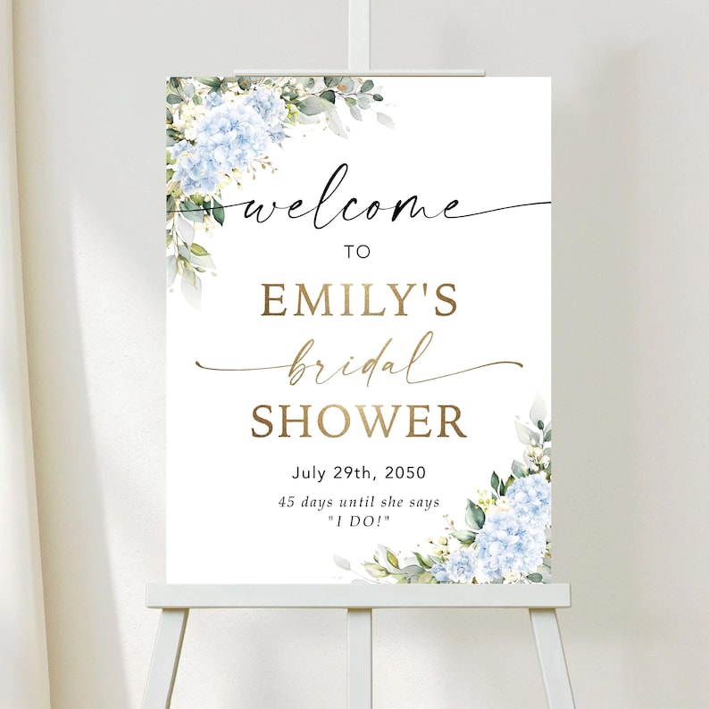 May include: A white sign on a wooden easel with the words "welcome to Emily's bridal shower" in gold and black script. The sign is decorated with blue floral accents and the date July 29th, 2050.