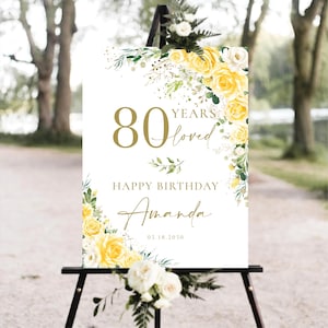 May include: A white sign on a black easel with the text "80 Years Loved Happy Birthday Amanda 05.18.2050" in gold script. The sign is decorated with yellow roses and greenery.