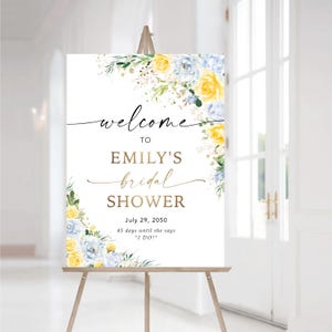 May include: A white sign on a wooden easel with floral accents. The sign reads "welcome to Emily's bridal shower" in elegant script. Yellow and blue flowers decorate the edges, with the date July 29, 2050, and a countdown.