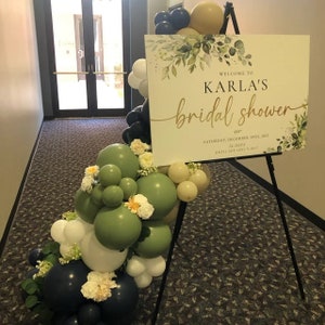 May include: A green, white, and navy blue balloon arch with white flowers and greenery. A sign with the text "WELCOME TO KARLA'S bridal shower" and the date "SATURDAY, DECEMBER 30TH, 2023" is on an easel.