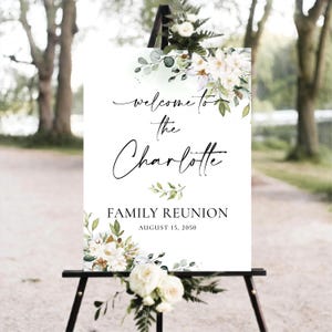 May include: A white sign on a black easel with floral accents. The sign reads "Welcome to the Charlotte Family Reunion August 15, 2050" in elegant script and serif fonts. White and green floral arrangements adorn the top and bottom.