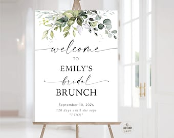 Op de afbeelding: Een wit bord met een groen aquarel bloemenmotief. De tekst luidt "Welkom bij Emily's bruidsbrunch, 10 september 2026, 120 dagen tot ze "Ja" zegt!"