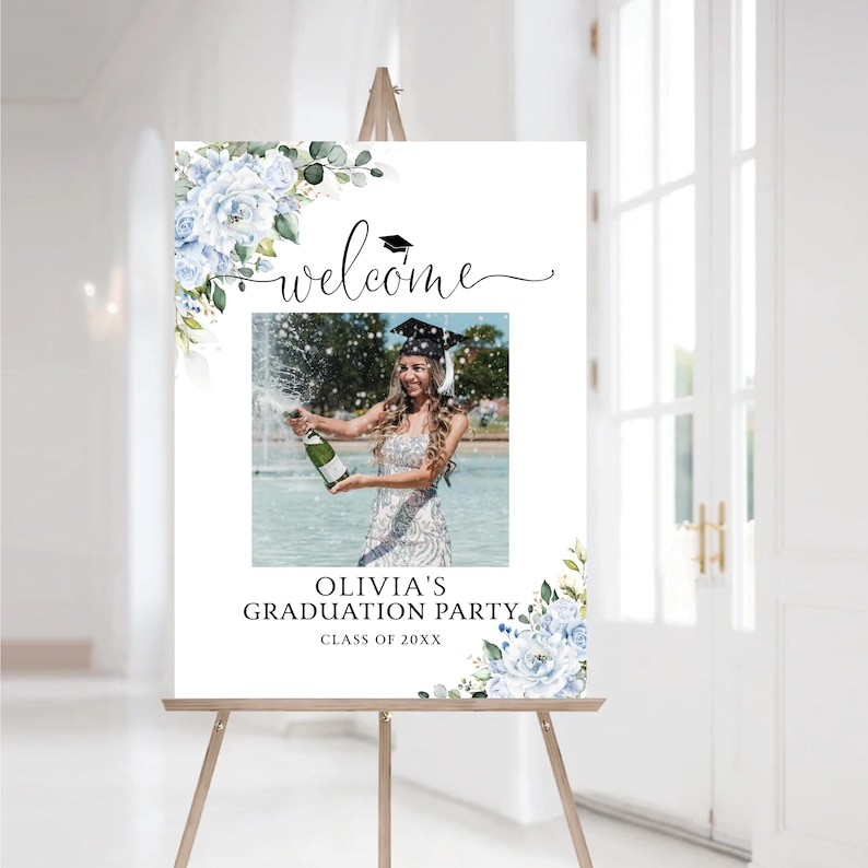 May include: A graduation party welcome sign on a wooden easel. The sign features a photo of a graduate popping champagne, with text that reads "Welcome" and "Olivia's Graduation Party, Class of 20XX." Floral accents in blue and green complete the design.