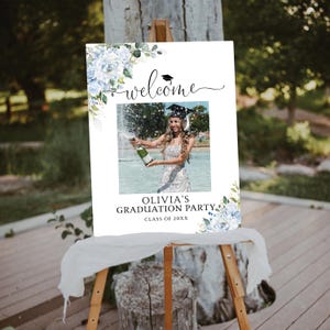 May include: A white graduation party welcome sign on an easel. The sign features a photo of a graduate popping champagne, with the text "welcome" and "Olivia's Graduation Party". Floral accents in blue and green complete the design.