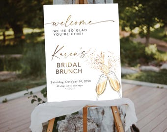 May include: A white sign with gold lettering announcing a bridal brunch for Karen. The sign features a watercolour illustration of champagne glasses with gold confetti. The text reads "Welcome, We're so glad you're here! Karen's Bridal Brunch, Saturday, October 14, 2050, 45 days until she says "I do!"