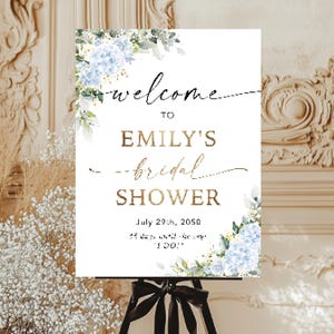 May include: A white sign for a bridal shower, with the words "Welcome to Emily's Bridal Shower" in gold and black script. The sign is decorated with blue floral accents and is displayed on a black easel.