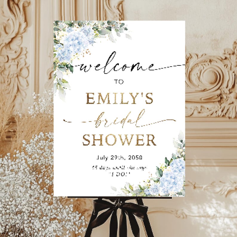 May include: A white sign for a bridal shower, with the words "Welcome to Emily's Bridal Shower" in gold and black script. The sign is decorated with blue floral accents and is displayed on a black easel.