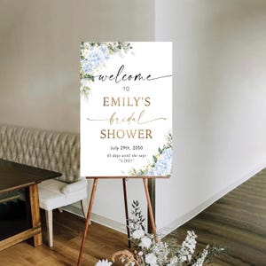 May include: A white sign on a wooden easel with the text "welcome to Emily's bridal shower." The sign is decorated with blue and green floral accents. The date July 29th, 2050, is displayed, along with the text "45 days until she says 'I DO!'"