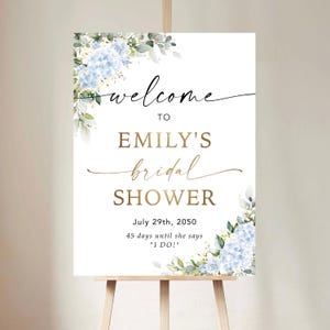 May include: A white sign on a wooden easel, announcing "welcome to Emily's bridal shower" in stylish script. The sign is adorned with blue hydrangea and foliage. The date July 29th, 2050, is shown, with the text "45 days until she says 'I DO!'"