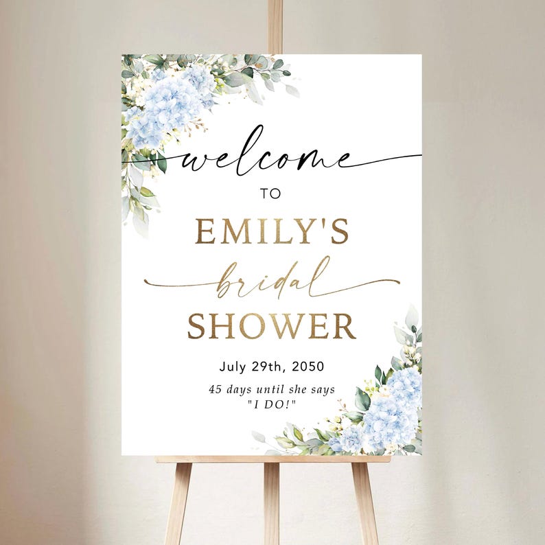 May include: A white sign on a wooden easel, announcing "welcome to Emily's bridal shower" in stylish script. The sign is adorned with blue hydrangea and foliage. The date July 29th, 2050, is shown, with the text "45 days until she says 'I DO!'"