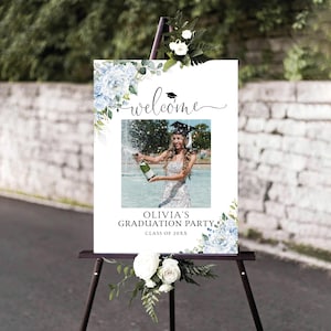 May include: A white graduation party welcome sign with blue floral accents and the text "welcome". The sign features a photo of a graduate popping champagne and reads "Olivia's Graduation Party, Class of 20XX". The sign is displayed on a black easel.