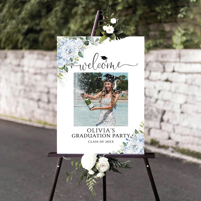 May include: A white graduation party welcome sign with blue floral accents and the text "welcome". The sign features a photo of a graduate popping champagne and reads "Olivia's Graduation Party, Class of 20XX". The sign is displayed on a black easel.