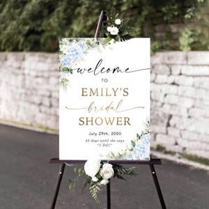 May include: A sign on a black easel, welcoming guests to Emily's bridal shower. The white sign features gold script and blue hydrangea floral arrangements. The date of the event, July 29th, 2050, is also displayed on the sign.