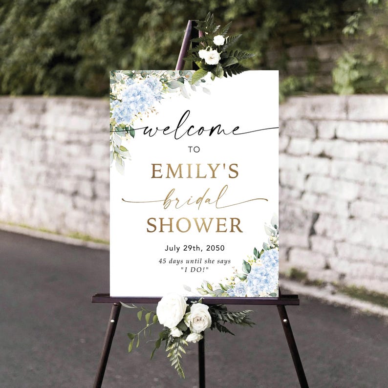 May include: A sign on a black easel, welcoming guests to Emily's bridal shower. The white sign features gold script and blue hydrangea floral arrangements. The date of the event, July 29th, 2050, is also displayed on the sign.