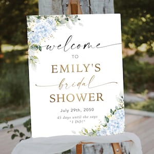 May include: A white sign with the words "welcome to EMILY'S bridal SHOWER" in gold and black script. The sign includes the date July 29th, 2050, and the text "45 days until she says 'I DO!'". The sign is decorated with blue and white floral accents.