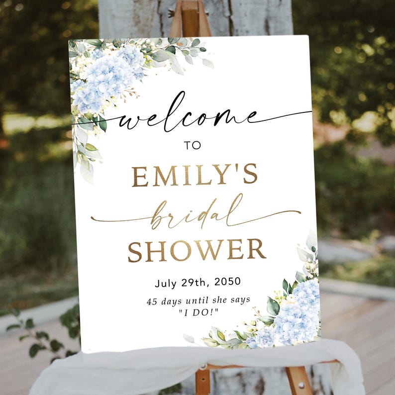 May include: A white sign with the words "welcome to EMILY'S bridal SHOWER" in gold and black script. The sign includes the date July 29th, 2050, and the text "45 days until she says 'I DO!'". The sign is decorated with blue and white floral accents.