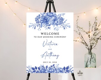 May include: A white wedding welcome sign with blue floral designs. The text reads "Welcome to our wedding ceremony Victoria and Anthony July 10, 2050".