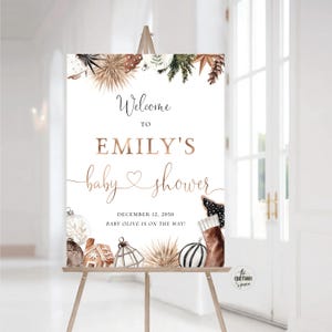 Puede incluir: Un cartel de bienvenida para la fiesta de bienvenida al bebé de Emily con un fondo blanco y un diseño de purpurina dorada. El cartel presenta un tema invernal con adornos, ramas de pino y una media. El texto dice "¡Bienvenido a la fiesta de bienvenida al bebé de Emily, 12 de diciembre de 2050, Baby Olive está en camino!"