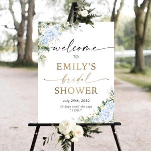 May include: A white sign on a black easel, adorned with blue hydrangea and foliage. The sign displays "welcome to Emily's bridal shower" in a stylish script, with the date July 29th, 2050, and the countdown.