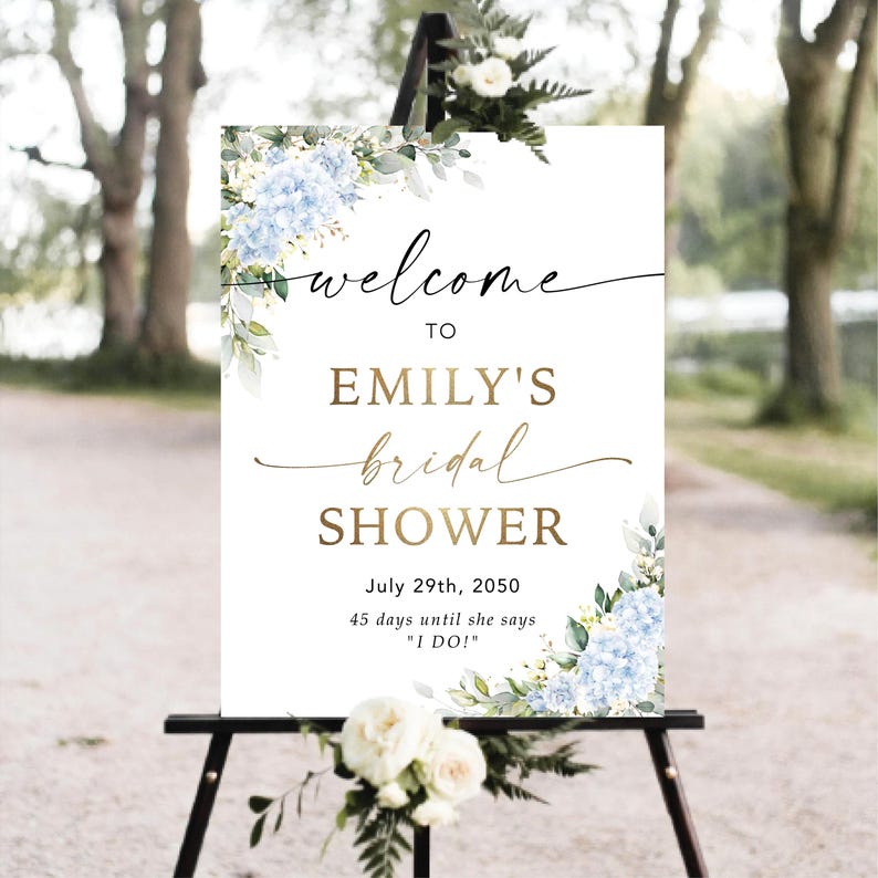May include: A white sign on a black easel, adorned with blue hydrangea and foliage. The sign displays "welcome to Emily's bridal shower" in a stylish script, with the date July 29th, 2050, and the countdown.