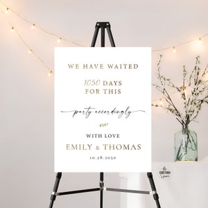 May include: White wedding sign with gold text that reads "We have waited 1050 days for this party accordingly" with a small green leaf design. The sign also includes the text "With love Emily & Thomas 10.28.2050".