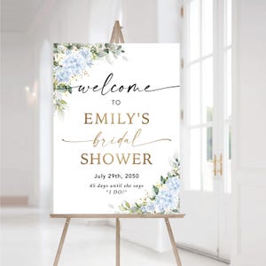 May include: A white sign on a wooden easel with the text "welcome to EMILY'S bridal SHOWER." The sign is decorated with blue hydrangea flowers and greenery. The date July 29th, 2050, and the phrase "45 days until she says 'I DO!'" are also included.