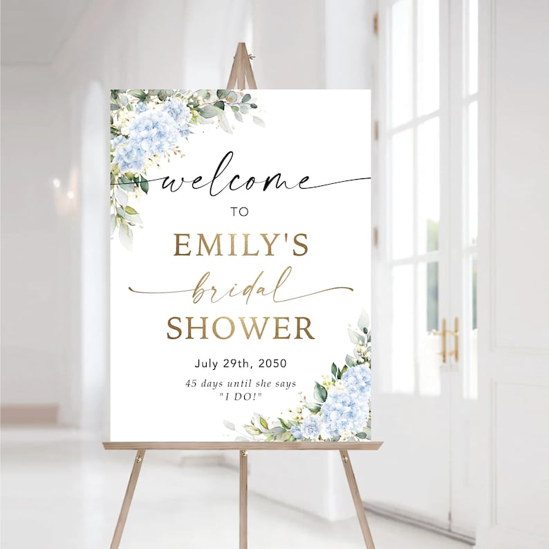 May include: A white sign on a wooden easel with the text "welcome to EMILY'S bridal SHOWER." The sign is decorated with blue hydrangea flowers and greenery. The date July 29th, 2050, and the phrase "45 days until she says 'I DO!'" are also included.
