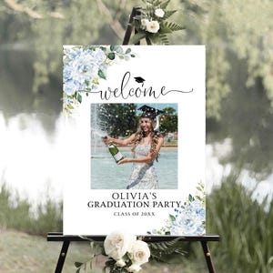 May include: A welcome sign for a graduation party displayed on a black easel. The sign has a photo of a graduate with champagne, the word "welcome", and details for "Olivia's Graduation Party Class of 20XX". It is decorated with blue floral designs.