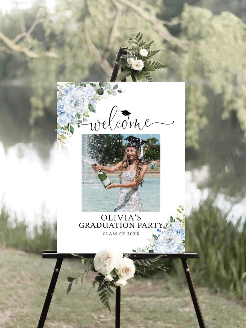 May include: A welcome sign for a graduation party displayed on a black easel. The sign has a photo of a graduate with champagne, the word "welcome", and details for "Olivia's Graduation Party Class of 20XX". It is decorated with blue floral designs.