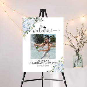 May include: A graduation party welcome sign on a black easel. The sign features a photo of a graduate popping champagne, surrounded by blue floral accents. The sign reads "welcome" and "Olivia's Graduation Party, Class of 20XX."