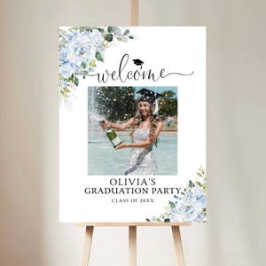May include: A graduation party welcome sign on an easel. The sign features a photo of a graduate popping champagne, surrounded by floral accents and the text "Welcome" and "Olivia's Graduation Party."