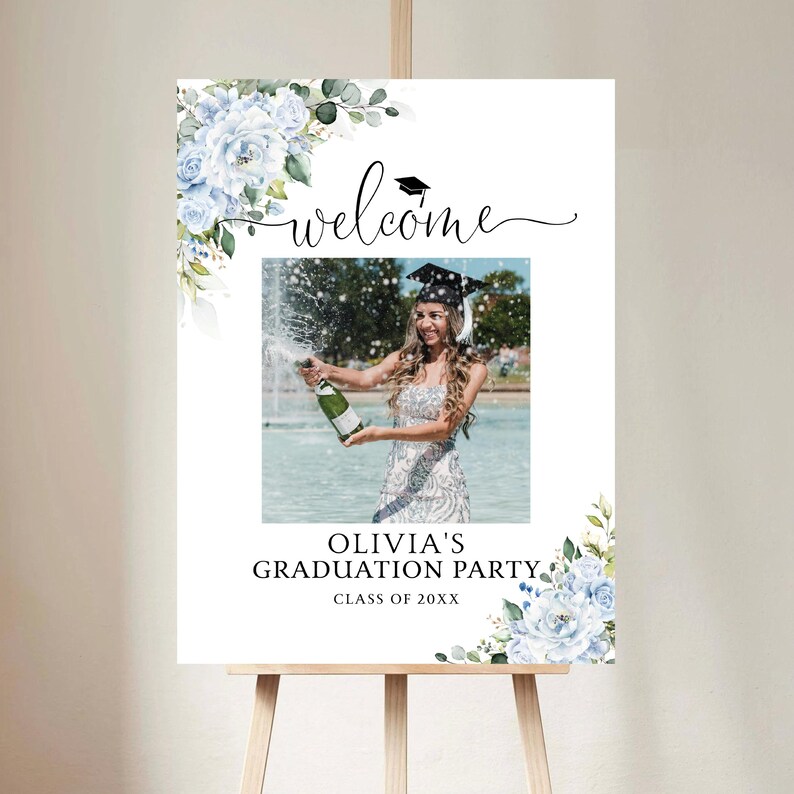 May include: A graduation party welcome sign on an easel. The sign features a photo of a graduate popping champagne, surrounded by floral accents and the text "Welcome" and "Olivia's Graduation Party."