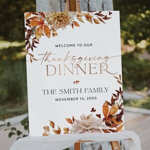 May include: A white sign with a watercolor floral border in shades of orange, brown, and cream. The text on the sign reads "Welcome to our Thanksgiving Dinner The Smith Family November 15, 2050".