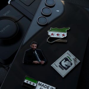 May include: Black smartphone with several enamel pins attached. Pins include a Syrian flag, a map of Syria, a postage stamp, a pin with a man's image, and a pin that says "MADE IN SYRIA".