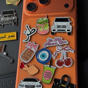 May include: An orange phone case adorned with various enamel pins. Pins include a green drink, a black handbag, a car, a pair of cherries, and text that says "In My Pilates Era".