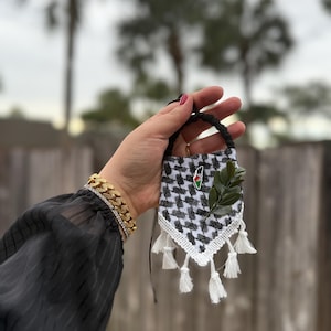 May include: A small, decorative black and white patterned textile with white tassels and a green leaf accent. The textile has a black cord loop and a pin with a flag design. The background is blurred, with palm trees and a wooden fence.