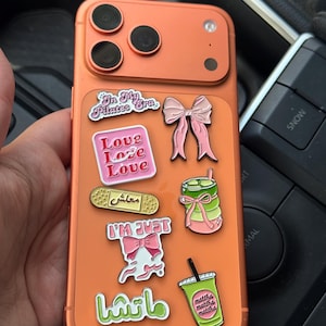 May include: An orange smartphone case decorated with various enamel pins, including a pink bow, a green matcha drink, a bandage with Arabic text, and text pins reading In My Pilates Era, Love Love Love, I'M JUST, and matcha.