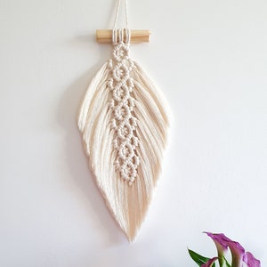 Puede incluir: Un tapiz de macramé blanco en forma de pluma con una varilla de madera en la parte superior. El tapiz está colgado en una pared blanca. Hay dos plantas en macetas y una gran roca de sal rosa del Himalaya en primer plano.