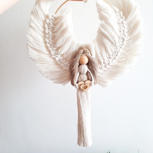 Op de afbeelding: Een witte macramé engel met een houten poppenhoofd en bruin haar, die een klein houten hartje vasthoudt. De engel hangt aan een gouden hoepel met een klein hartje.