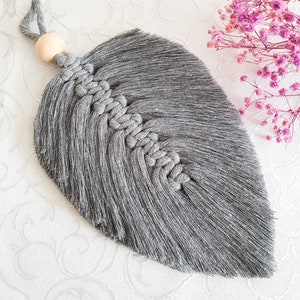 May include: A grey macrame feather wall hanging with a wooden bead at the top. The feather is made with a thick, soft yarn and has a fringed edge.