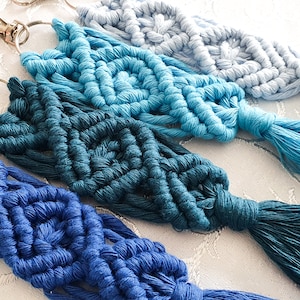 May include: Four macrame keyrings in varying shades of blue, from dark indigo to pale sky blue. Each keyring has a woven pattern, a tassel, and a silver-coloured clasp. The keyrings are displayed on a white surface.
