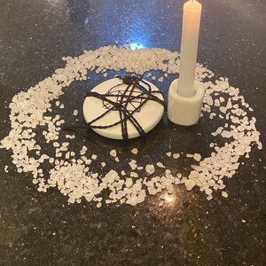 Puede incluir: Una vela blanca con una llama azul se encuentra en un portavelas blanco sobre una superficie negra. Un círculo de sal blanca rodea la vela y un disco de cerámica blanca con una cuerda negra atada alrededor.