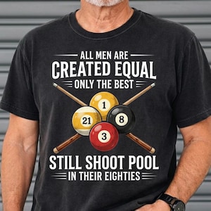 May include: Black t-shirt with a pool-themed graphic. The design includes crossed pool cues, four billiard balls numbered 1, 3, 8, and 21, and the text "ALL MEN ARE CREATED EQUAL ONLY THE BEST STILL SHOOT POOL IN THEIR EIGHTIES."