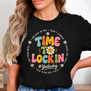 May include: Black t-shirt featuring the text "TIME TO LOCK IN" in vibrant colours, with a smiling flower graphic. The design includes the hashtag "Yesterday" and the phrases "I believe in you", "Don't stress", "Do your best", and "Rock the test".