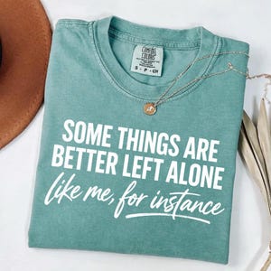 May include: A teal Comfort Colors t-shirt with the text "SOME THINGS ARE BETTER LEFT ALONE like me, for instance" in white. A silver necklace with a small pendant rests on the shirt. The shirt is folded on a white surface.