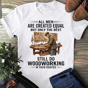 Op de afbeelding: Wit T-shirt met de tekst "ALL MEN ARE CREATED EQUAL BUT ONLY THE BEST STILL DO WOODWORKING IN THEIR EIGHTIES." De afbeelding toont een houtbewerker aan een werkbank. Bruine leren schoenen en een blauwe jeans zijn ook zichtbaar.