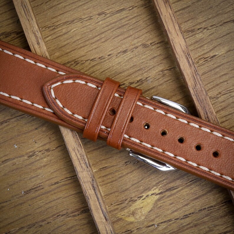 May include: Close-up of a brown leather watch strap with white stitching. The strap features a buckle and several adjustment holes. The leather has a smooth texture and is set against a wooden background.