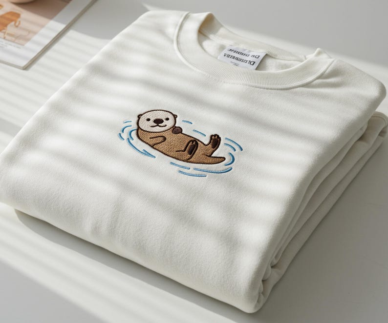 Pode incluir: Camisola creme com uma lontra bordada flutuando em &aacute;gua azul. A lontra &eacute; marrom com o rosto branco e est&aacute; deitada de costas. A camisola est&aacute; dobrada cuidadosamente sobre uma superf&iacute;cie branca.